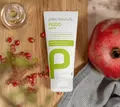 En hvid tube med teksten "peclavus PODO care FUSSLOTION GRANATAPFEL POMEGRANATE FOOT LOTION HAUTREGENERATION SKIN REGENERATION" ligger på et træbord omgivet af granatæblefrø, en granatæblefrugt og en grå klud.