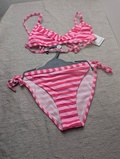 Et pink- og hvidstribet bikinisæt med flæseoverdel og bindeunderdel er udstillet på en lysegrå flade, hvor underdelen hænger på en plastikbøjle, og overdelen er lagt fladt over den.