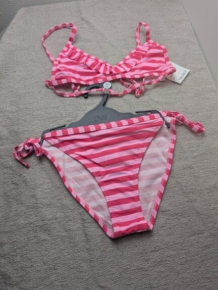 Et pink- og hvidstribet bikinisæt med flæseoverdel og bindeunderdel er udstillet på en lysegrå flade, hvor underdelen hænger på en plastikbøjle, og overdelen er lagt fladt over den.