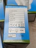 Nærbillede af en pære-emballage, der viser specifikationer: 230V/50Hz, E27, 99 Ra, 122 mA, 2000H levetid, varm hvid 2700K, 0,5 sek forsinkelse. Indeholder ikoner til certificeringer og en hjemmeside-URL. Emballagen er blå og grøn.