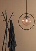 En moderne pendel med rund glasskærm hænger ved siden af en minimalistisk knagerække. En mørk jakke er draperet over en af krogene. Baggrunden er en almindelig, lysebrun væg.
