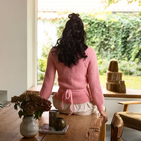 Ryggen af model sidder på et bord iført strikket cardigan Embrace fra filcolana i Vilja og Tilia i Lyserød 370 Flamingo