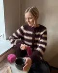 PetiteKnit iført Key Cardigan fra PetiteKnit i striber, sidder ved vindue med en kop kaffe og strikketøj.