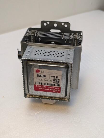 En metalmikrobølgetransformator mærket "LG 2M286" med en advarsel, der lyder "FARE High Voltage Microwave Protector." Komponenten er sølv med perforeringer og monteringsbeslag på toppen, placeret på en hvid overflade.