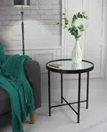 Et rundt sidebord med glasplade med sort metalstel står ved siden af en grå sofa draperet med et blågrønt tæppe. En hvid vase med grønt løv og en smartphone hviler på bordet. Baggrunden har en hvid murstensvæg og gulvlampe.