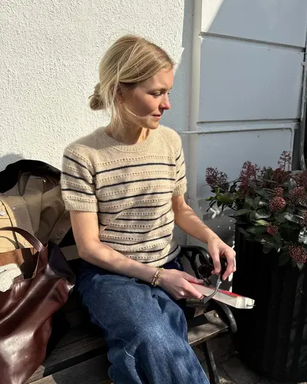 Ida Tee fra PetiteKnit i beige og mørkeblå striber, kvinde sidder udendørs på bænk med bog og lædertaske ved siden af.