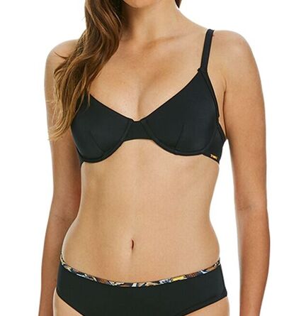 En kvinde iført sort bh og matchende sorte trusser med mønstret linning står på en almindelig hvid baggrund. Hendes ansigt er delvist ude af billedet.