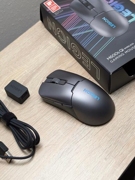 Trådløs gaming mus med LEGION branding sidder på en træoverflade. Ved siden af er der et USB-kabel og en lille adapter. Museboksen, der har et farverigt design og produktdetaljer, er delvist synlig i baggrunden.