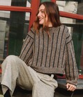 Person iført Edda Sweater af Hanne Rimmen i farverne Charcoal som bundfarve og Oak/Stone som mønsterfarver sidder afslappet foran en bygning med farvede vinduesrammer.