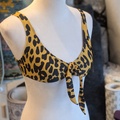 En mannequin viser en gul og sort bikinitop med leopardprint og bindebånd foran. Baggrunden viser stofruller og en struktureret væg.