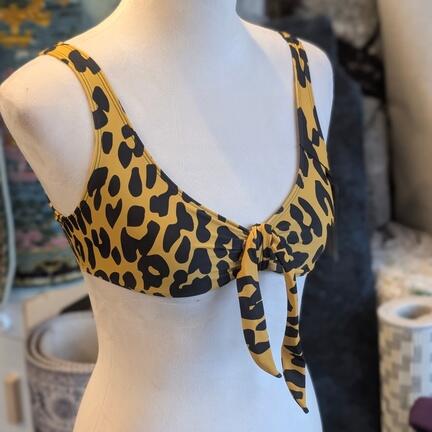 En mannequin viser en gul og sort bikinitop med leopardprint og bindebånd foran. Baggrunden viser stofruller og en struktureret væg.