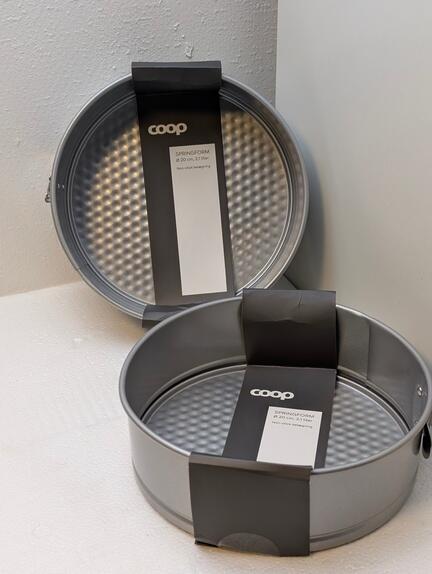 To runde, grå non-stick springforme udstillet på en hvid overflade, hver med en sort etiket fra "Coop". Den ene gryde står oprejst, og den anden er vippet, hvilket viser dens strukturerede indre overflade.