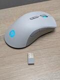 En hvid trådløs computermus med sideknapper og et lysende blåt logo sidder på en træoverflade. Foran den er der en lille USB-modtager.