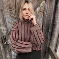 Person iført Edda Sweater designet af Hanne Rimmen i farverne Wine som bundfarve og Oak/Stone som mønsterfarver står foran en rustik trædør med armene krydset.