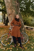 Andrea Hood balaclava i mørk brun fra Isager, båret over en lang camel-frakke og belte, stående op ad et træ med en rød cykel i et parklandskab med efterårsløv.