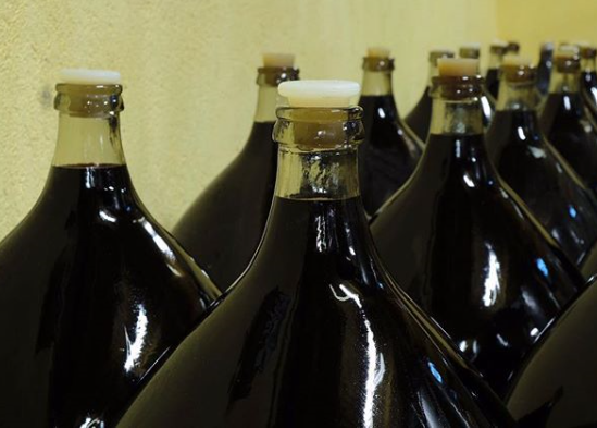 Store runde glasflasker fyldt med rødvin. Der er prop på flaskerne. Glasflaskerne står på en lang række og anvendes til lagring af rødvinen