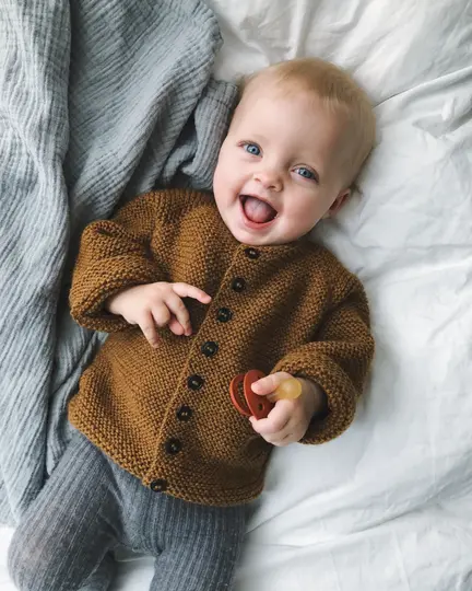 baby på seng ifoert begynderjakken fra petiteknit i lysebrun