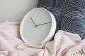 Et hvidt analogt ur med minimalistisk design og ingen tal er støttet mod en marineblå pude med geometriske mønstre. Den hviler på et blødt, lyserødt tæppe. Indstillingen antyder et hyggeligt, moderne interiør.