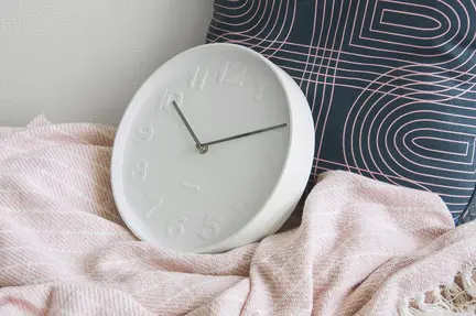 Et hvidt analogt ur med minimalistisk design og ingen tal er støttet mod en marineblå pude med geometriske mønstre. Den hviler på et blødt, lyserødt tæppe. Indstillingen antyder et hyggeligt, moderne interiør.