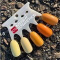 En plastik prøveholder med fem kunstige negle i forskellige nuancer af gul og orange. Disse nuancer er mærket: CANDLELIGHT, CYPSE, ALONG IN MARIGOLD, HAZELLE MAREY, JELLIED. Tekst: ICG INSIGHT COSMETICS. Baggrunden er grus.