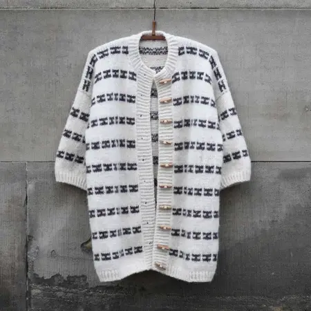 Karla Cape strikket i hvid bund med mørkt mønster, hængende på bøjle.