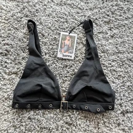 En sort bralette med øskendetaljer og en justerbar strop vises på et gråt tekstureret tæppe med et påsat produktmærke.