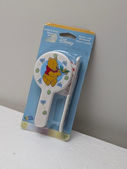 Et pakket Disney Plys-kam- og børstesæt til babyer, med en illustration af Plys siddende med en rød skjorte, omgivet af grønne blade og blå sommerfugle på børsten.