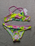 En todelt bikini med pink kant og et blomstermønster i pink, grøn og gul, der ligger fladt på en lysegrå struktureret overflade. Der sidder et mærke på bikinioverdelen.