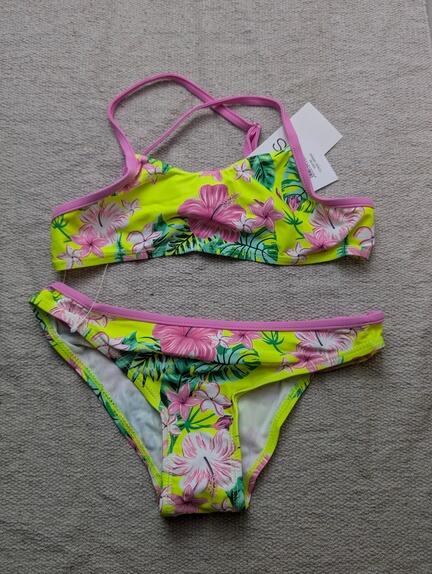 En todelt bikini med pink kant og et blomstermønster i pink, grøn og gul, der ligger fladt på en lysegrå struktureret overflade. Der sidder et mærke på bikinioverdelen.
