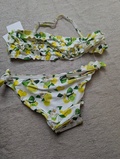 Et todelt bikinisæt med hvid baggrund og citronprint med grønne blade og gule citroner. Overdelen har flæsedetaljer, og underdelen har bindebånd i siden. Begge dele er lagt ud på en lys overflade.