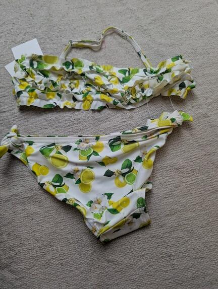 Et todelt bikinisæt med hvid baggrund og citronprint med grønne blade og gule citroner. Overdelen har flæsedetaljer, og underdelen har bindebånd i siden. Begge dele er lagt ud på en lys overflade.