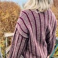 Bagfra visning af Edda Sweater af Hanne Rimmen, strikket i Wine og Oak/Stone. Personen står ved en sø omgivet af siv og efterårsfarver.